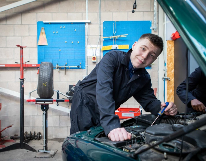 Student in a black overall smiling while working on a car engine, with a workshop setup in the background. Student in a black overall smiling while working on a car engine, with a workshop setup in the background.